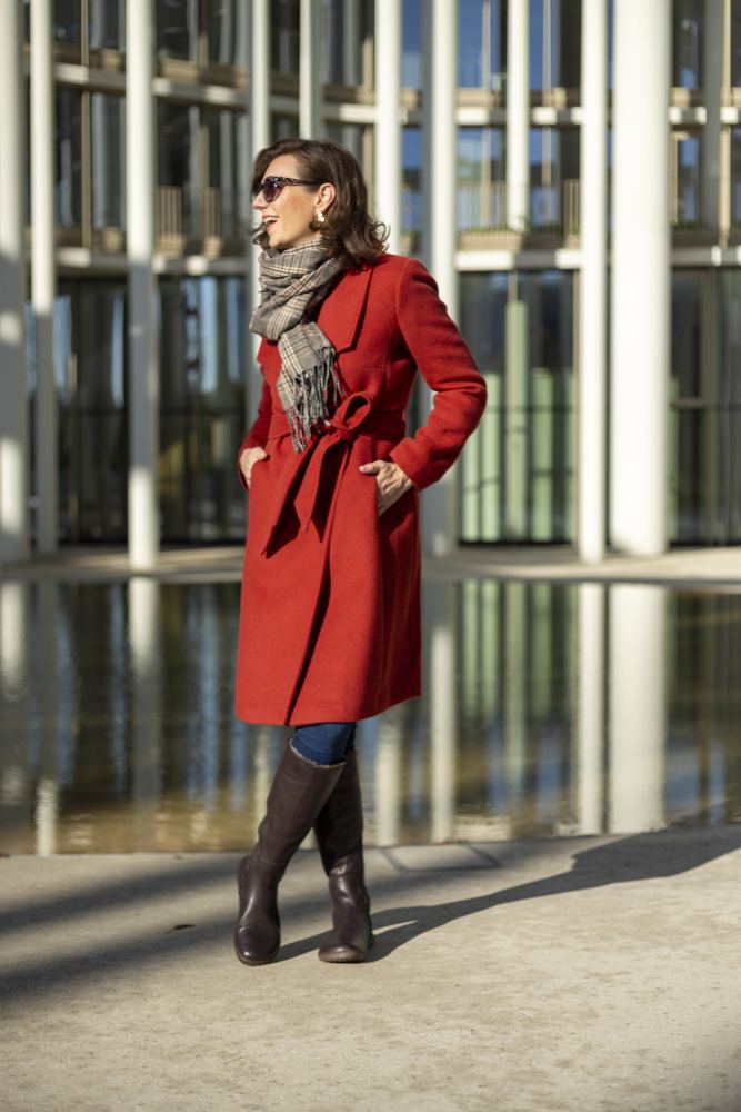 Eine Person in rotem Mantel, grauem Schal und Sonnenbrille steht selbstbewusst vor einem modernen Glasgebäude mit Säulen. Ihre QATCH Winter Brown Boots von ZAQQ aus luxuriösem Nappaleder schimmern dezent, während das Sonnenlicht Schatten auf den Boden wirft.