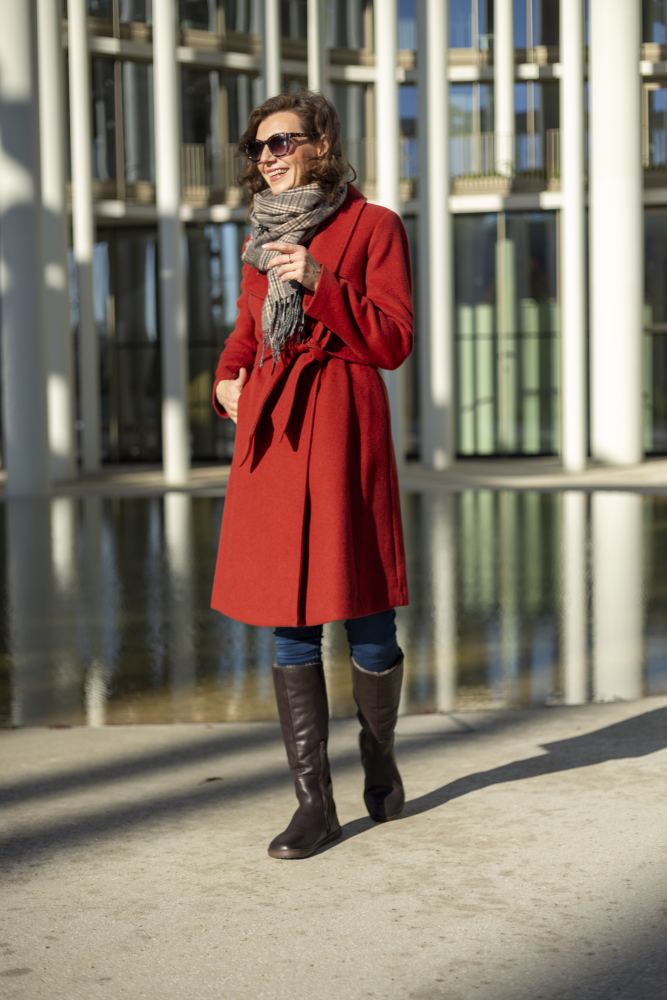 Eine Person in rotem Mantel, Sonnenbrille und Schal geht selbstbewusst an einer reflektierenden Oberfläche vorbei, im Hintergrund ein modernes Gebäude. Sie kombiniert ihre Jeans mit hohen ZAQQ QATCH Winter Brown-Stiefeln. Die gut beleuchtete Szene wird durch das warme Sonnenlicht noch verstärkt.