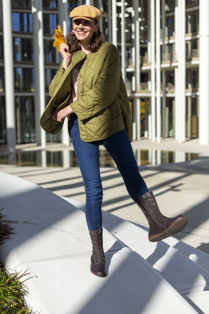 Eine Frau in einer grünen Steppjacke und Schiebermütze hebt verspielt ein Bein und hält dabei Herbstblätter in der Hand. Sie steht draußen in einem sonnenbeschienenen Bereich mit einem modernen Glasgebäude im Hintergrund und trägt Jeans und braune ATTRAQT Stretch Nappa-Schuhe von ZAQQ.