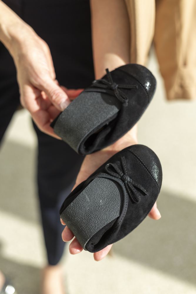 Eine Person hält ein Paar TWIST Velours Black Ballerinas von ZAQQ, aus schwarzem Veloursleder mit hübschen Schleifen an den Zehen. Diese flexiblen und leichten Schuhe sind so konzipiert, dass sie sich leicht zusammenfalten lassen und ein Barfußgefühl vermitteln. Der Hintergrund ist eine helle, verschwommene Oberfläche.