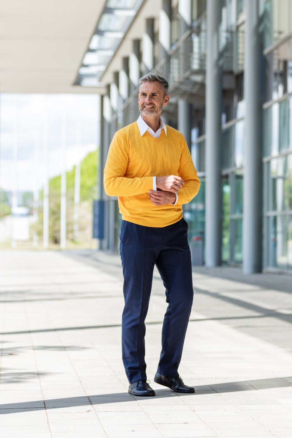 Ein Mann in gelbem Pullover und dunkler Hose steht vor einem modernen Gebäude mit großen Fenstern und rückt seinen Ärmel zurecht. Selbstbewusst schreitet er in seinen schwarzen ZAQQ ARQ-Stiefeln den sonnenbeschienenen Weg entlang, während Bäume und ein blauer Himmel eine ruhige Kulisse bilden.