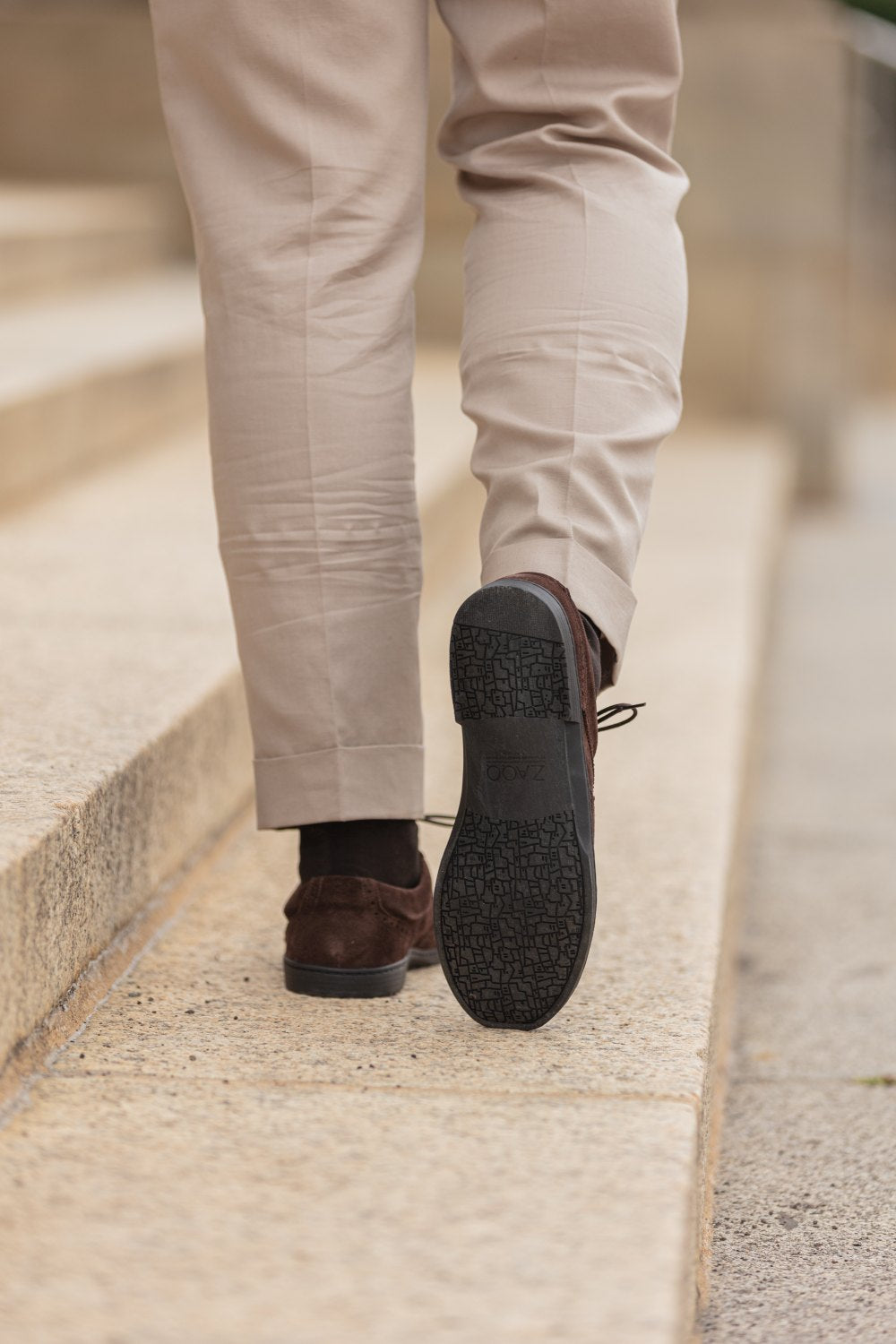 Eine Person in beigen Hosen läuft in einem Paar BRIQ Brogue Velours Brown-Schuhen von ZAQQ aus französischem Kalbleder Steinstufen hinauf. Die Sohle des linken Schuhs ist sichtbar und weist ein strukturiertes Muster auf.