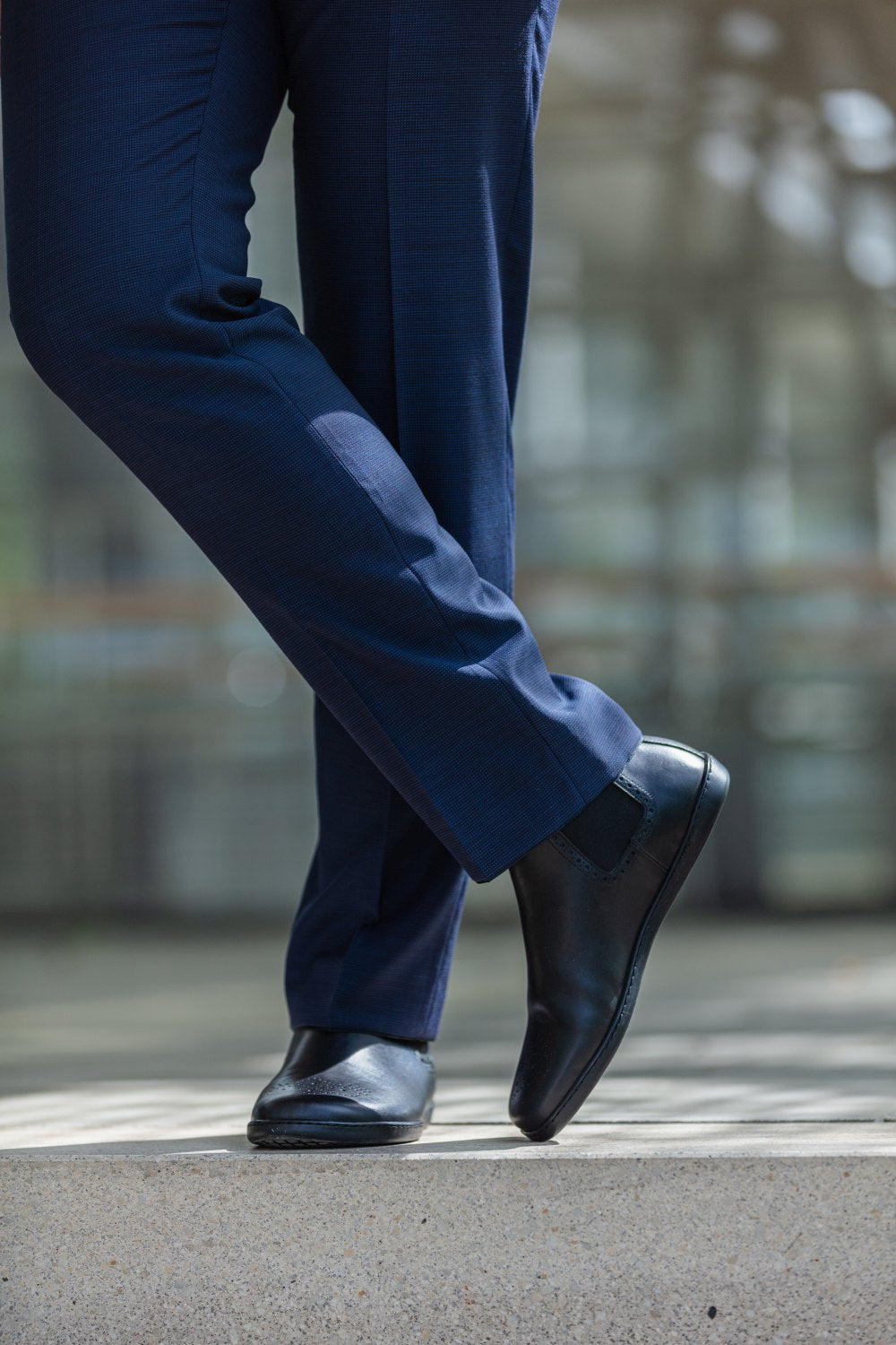 Eine Person in dunkelblauer Anzughose steht im Freien und schlägt die Beine übereinander. Dabei präsentiert sie den EQUITY Brogue Black von ZAQQ. Die Chelsea Boots aus hochwertigem Nappaleder, die sie trägt, ergänzen ihren stylischen Look. Im unscharfen Hintergrund fällt ein modernes Glasgebäude auf.