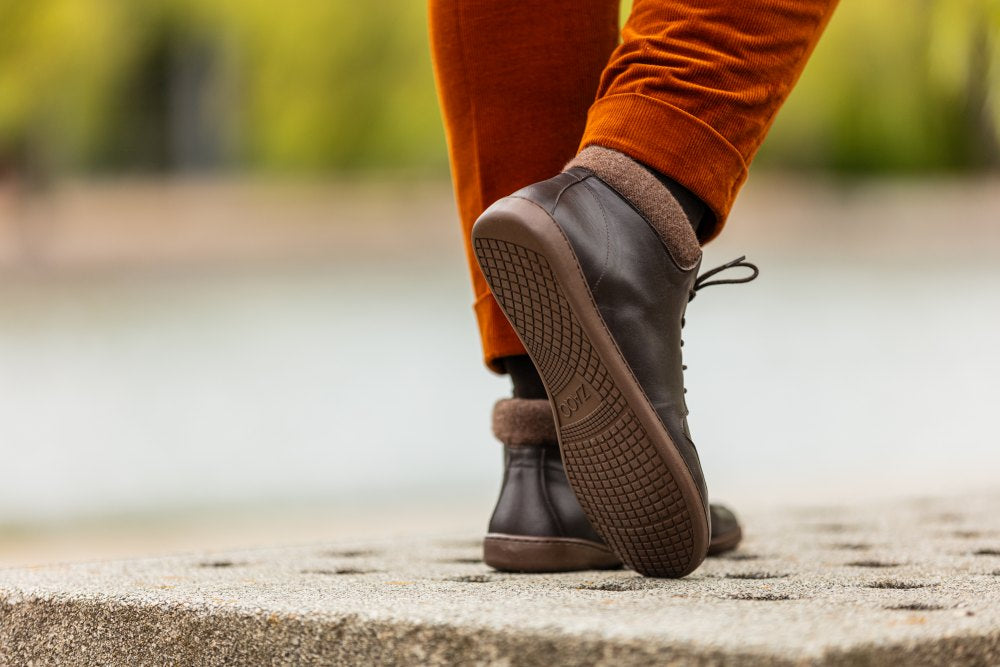 Nahaufnahme einer Person in orangefarbenen Hosen und braunen ZAQQ NEWQUAY-Schuhen, die auf einem Steinweg läuft. Der Fokus liegt auf dem Schuhwerk, der Hintergrund ist leicht unscharf.