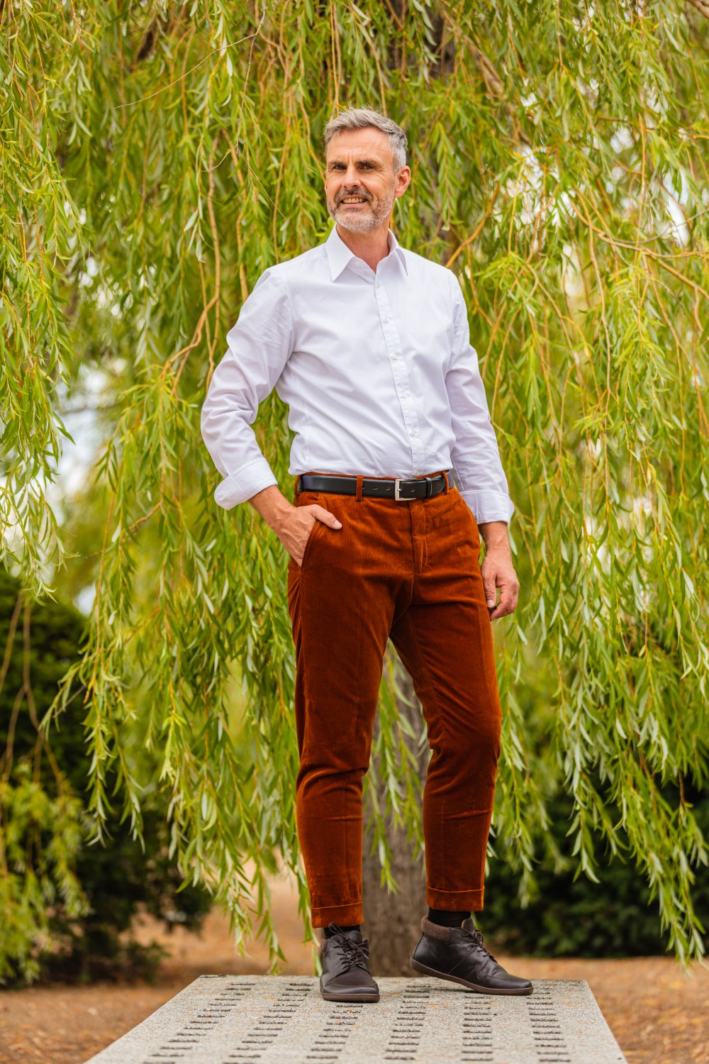 Ein Mann mit grauem Haar und Bart steht selbstbewusst da, trägt ein weißes Hemd und eine braune Hose. Er steht im Freien vor herabhängenden grünen Weidenzweigen, eine Hand in der Tasche und in der anderen präsentiert er stilvolles NEWQUAY Brown von ZAQQ, gefertigt aus luxuriösem Nappaleder.