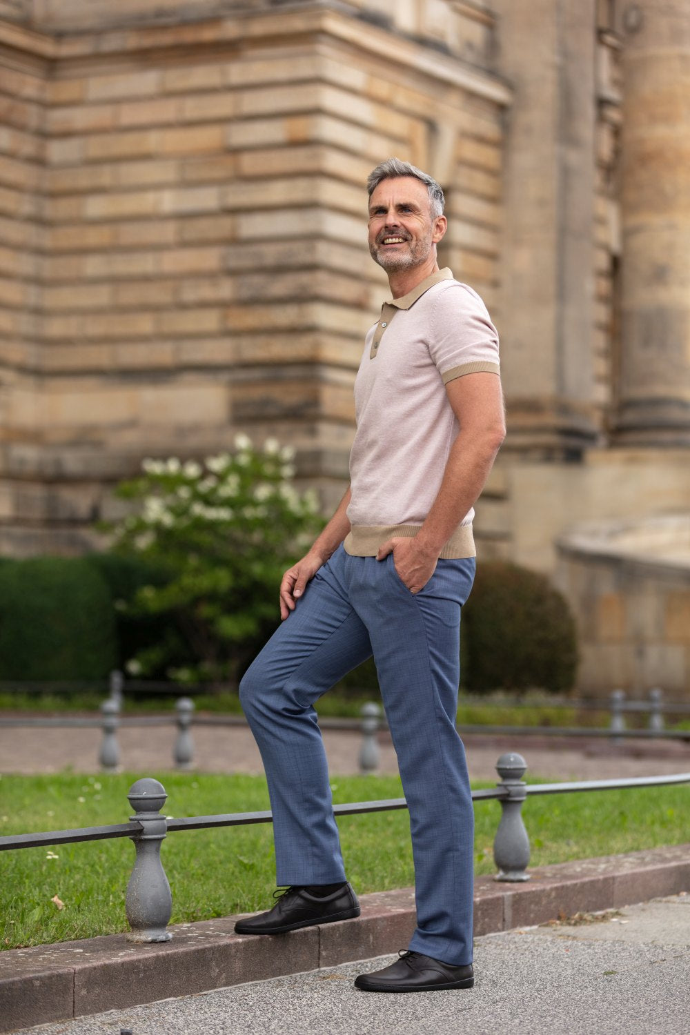 Ein Mann steht im Freien neben einem historischen Steingebäude und lächelt in seiner Business-Casual-Kleidung: ein helles Poloshirt, blaue Hosen und dunkelbraune ZAQQ PEAQ-Schuhe. Mit den Händen in den Taschen und einem Fuß auf einem niedrigen Zaun ruht er vor der Kulisse üppigen Grüns entspanntes Selbstvertrauen.