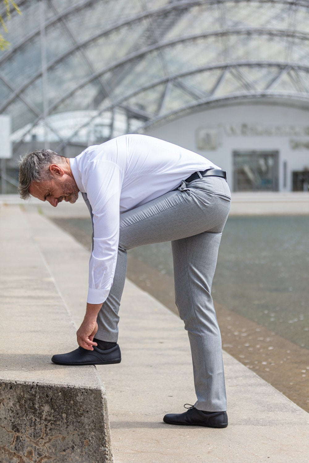 Ein Mann in weißem Hemd und grauer Hose schnürt seine stylischen PEAQ Vegan Black mit atmungsaktivem Mikrofaserfutter und stellt einen Fuß auf einen Betonvorsprung. Er steht im Freien neben einem Wasserspiel mit einer modernen Glaskuppelstruktur im Hintergrund. Die Schuhe sind Teil der Kollektion der Marke ZAQQ.