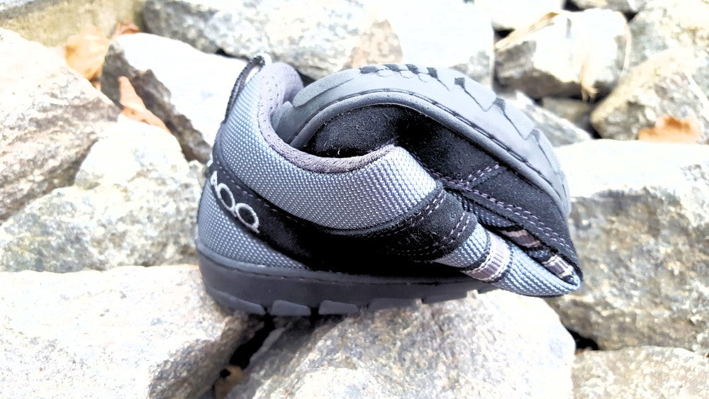 Ein kleiner ZAQQ SEEQ Black-Schuh in schwarz-grauem Design mit Riemen und offener Ferse steht auf einem Haufen grauer Steine. Dieser Barfußschuh wurde für Outdoor-Abenteuer entworfen und verfügt über ein gesticktes Logo, das seine Kombination aus Stil und Funktionalität unterstreicht.