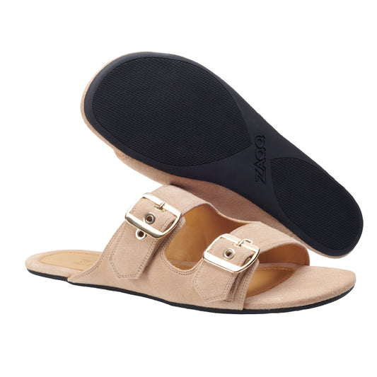 Die UQO Taupe Slide-Sandalen von ZAQQ aus Veloursleder in Beige haben zwei goldene Schnallen. Eine Sandale ist flach, während die andere abgewinkelt ist, um ihre schwarze strukturierte Sohle zu präsentieren. Mit ihrem minimalistischen Design bieten diese Schuhe sowohl Stil als auch Vielseitigkeit.