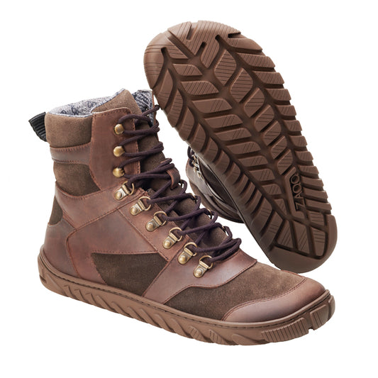 Die braunen EXPLORER Waterproof-Stiefel von ZAQQ sind hohe Lederschuhe mit dunklen Schnürsenkeln und goldenen Ösen. Sie sind ideal für Outdoor-Abenteuer und haben eine robuste, strukturierte Sohle für hervorragenden Halt. Ein Stiefel ist aufrecht abgebildet, um das Seitenprofil zu zeigen, während der andere angewinkelt ist, um das Sohlendesign hervorzuheben.