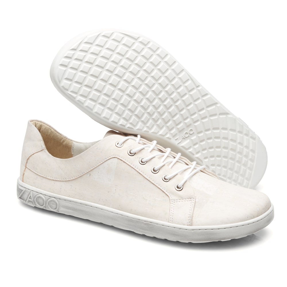 Die QORE Low Vegan White Sneaker von ZAQQ haben sichtbare weiße Schnürsenkel und eine strukturierte Sohle. Ein Schuh steht aufrecht, während der andere geneigt ist, um das Muster der Sohle zu zeigen. Ein dezentes Logo an der Seite unterstreicht ihr schlichtes Design.