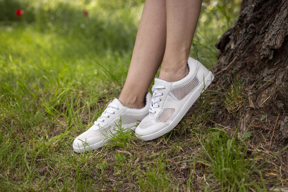 Eine Person lehnt an einem Baum, ihre weißen ZAQQ ALIQANTE-Sneakers ruhen auf dem sonnenbeschienenen Gras. Nur Beine und Füße sind zu sehen, umgeben von grünem Laub und einem robusten Baumstamm, was die Balance zwischen Stil und Atmungsaktivität in dieser ruhigen Parklandschaft unterstreicht.