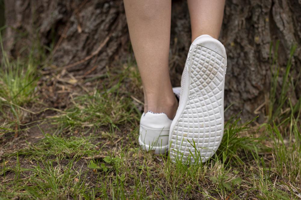 Eine Person, die die eleganten ALIQANTE White Sneaker von ZAQQ trägt, lehnt an einem Baumstamm. Ein Fuß steht flach auf dem Boden, während der andere angewinkelt ist, sodass die atmungsaktive Sohle sichtbar ist. Die Schuhe heben sich vom mit Gras und Erde bedeckten Boden ab.