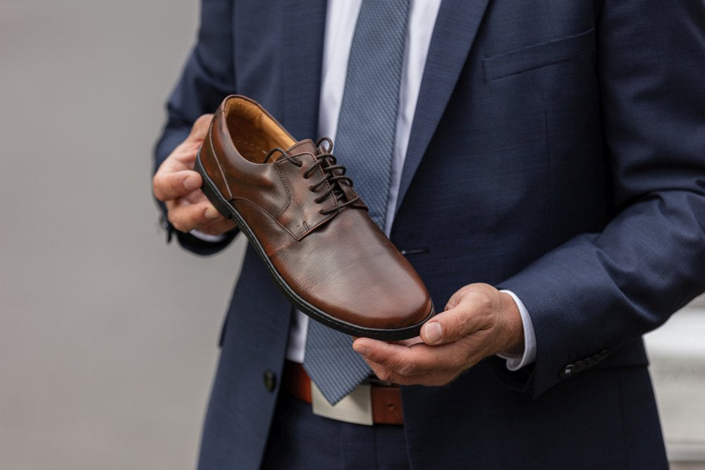 Eine Person in marineblauem Anzug und Krawatte hält einen BRIQ Antique Brown von ZAQQ und präsentiert dessen Design. Der aus feinem Kalbleder gefertigte Schuh sieht vor dem verschwommenen Hintergrund besonders elegant aus.