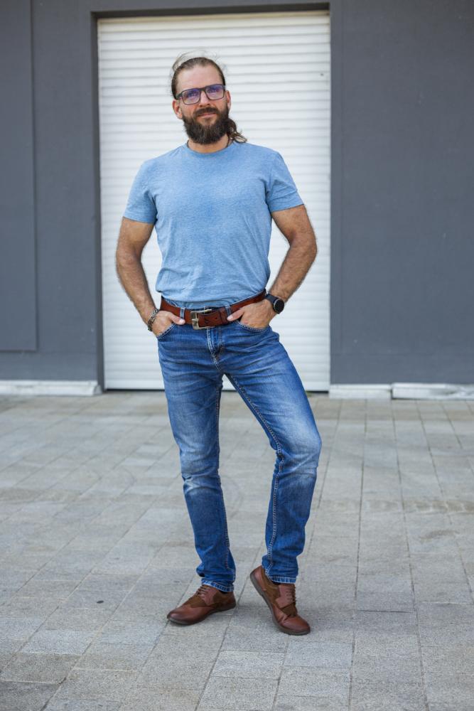 Ein bärtiger Mann mit langen Haaren und Brille, der ein blaues T-Shirt trägt, steht selbstbewusst mit den Händen in den Hüften. Er trägt Jeans und braune ZAQQ BRIQ Brogue Antique Cognac-Schuhe und posiert vor einem grauen Gebäude mit einer Tür mit Fensterläden.