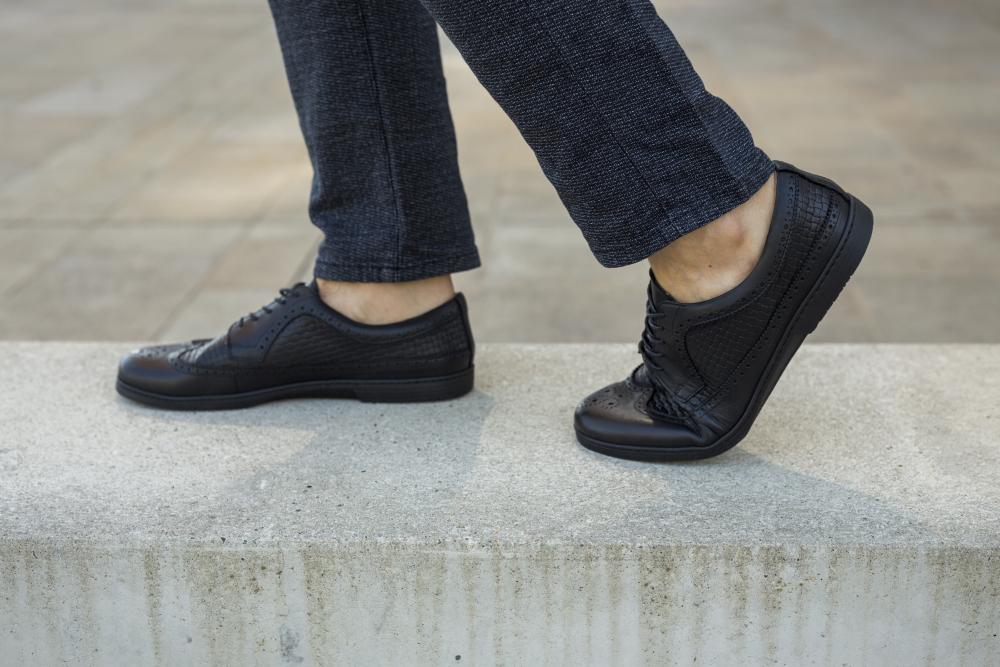 Eine Person läuft auf einem Betonvorsprung und trägt elegante ZAQQ BRIQ Brogue Tresse Black-Schuhe und dunkle Hosen. Der Außengehweg im Hintergrund verstärkt das elegante Ambiente der Szene.