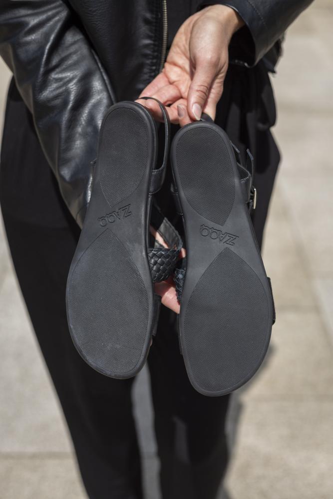 Eine Person in einer schwarzen Lederjacke hält ein Paar feminine schwarze Sandalen hoch und präsentiert auf den Sohlen den eingeprägten Markennamen ZAQQ. Das schlichte Design in BROOQE Black hebt sich vom hellgrauen Pflaster im Hintergrund ab.