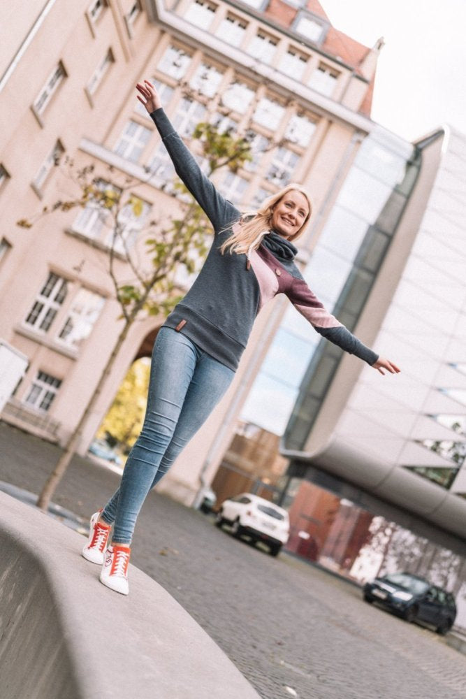 Eine Frau balanciert verspielt mit ausgestreckten Armen auf einer niedrigen Mauer. Sie trägt Jeans, einen Pullover und leuchtend rote Barfuß-Sneakers mit Kautschuksohle von ZAQQ CEDRIQ. Im Hintergrund sind moderne und ältere Gebäude zu sehen, und auf der Kopfsteinpflasterstraße parken ein paar Autos.