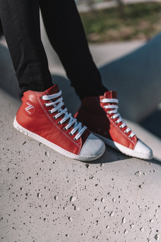 Eine Person steht selbstbewusst auf einer strukturierten Betonoberfläche und trägt CHUQQS Red von ZAQQ – stylische rote High-Top-Sneaker aus Nappaleder mit weißen Schnürsenkeln und Sohlen. Diese Sneaker, die an der Seite mit einem runden Logo verziert sind, ergänzen schwarze Hosen perfekt für einen eleganten Look.