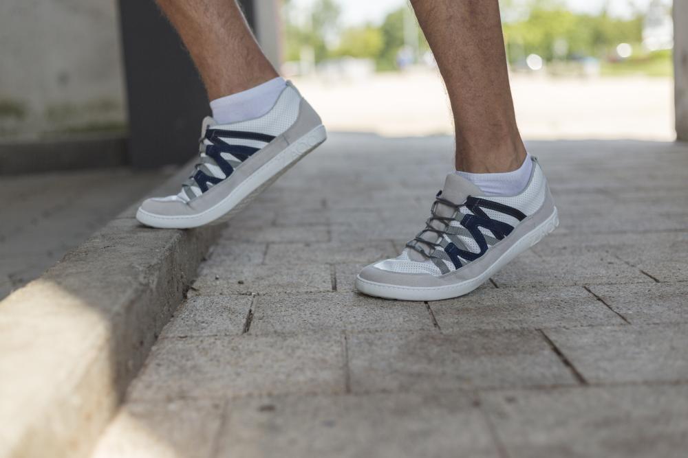 In den DRAQE White Grey Sneakers von ZAQQ mit schwarzen Schnürsenkeln tritt eine Person auf einen Bordstein entlang eines sonnenbeschienenen, mit Ziegeln gepflasterten Gehwegs. Diese Schuhe aus hochwertigem Nappaleder verschmelzen mühelos mit dem verschwommenen Grün dahinter.