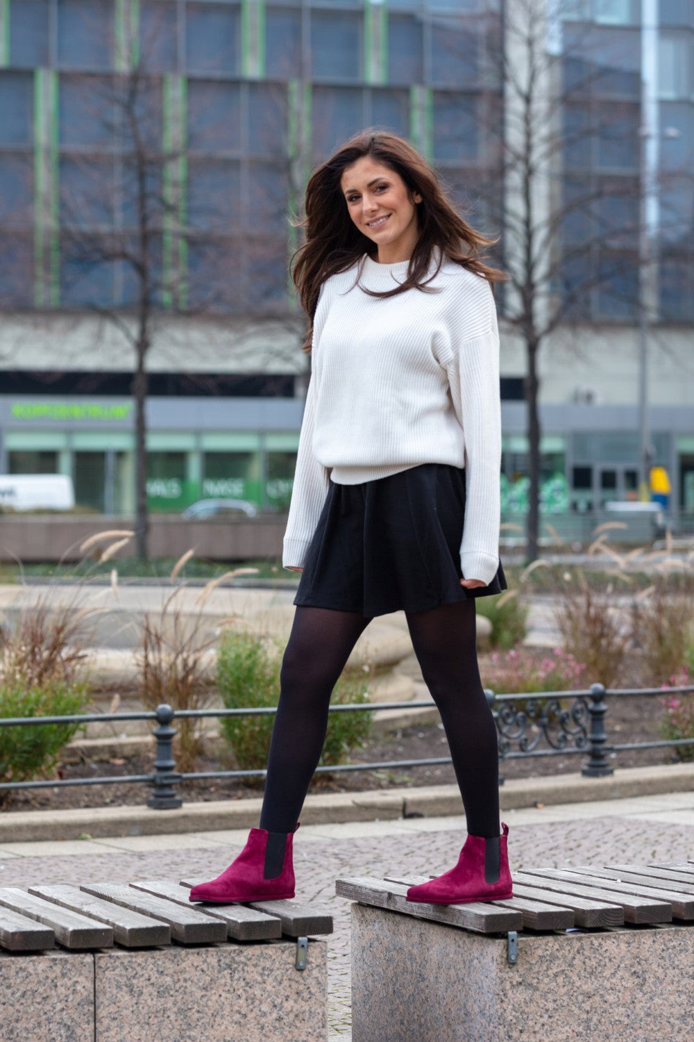 Eine Frau mit langen braunen Haaren steht lächelnd auf einer Steinbank im Freien. Sie trägt einen weißen Pullover, einen schwarzen Rock, schwarze Strumpfhosen und auffällige rote EQUITY Berry-Stiefel von ZAQQ. Im Hintergrund sind Gebäude und eine Parkanlage zu sehen.