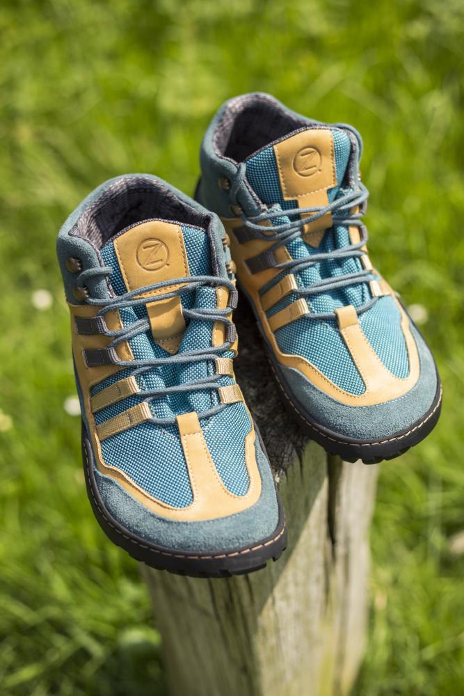 Ein Paar ESQAPE Blue Orange Waterproof Wanderschuhe von ZAQQ, mit einem auffälligen blau-beigen Farbschema mit schwarzen Sohlen und dem unverwechselbaren ZAQQ TERRA Sohlendesign, ruht auf einem Holzpfosten. Diese wasserdichten Schuhe heben sich vor einem Hintergrund aus grünem Gras ab und vereinen Stil und Funktionalität mit ihren mehreren Riemen für jedes Abenteuer.