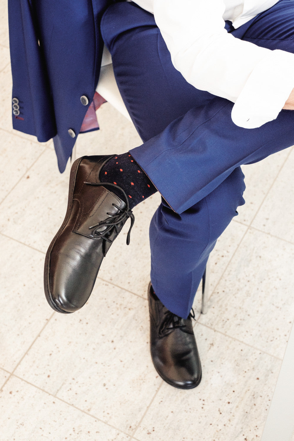 Eine Person in blauer Hose, weißem Hemd und blauem Blazer sitzt mit übereinandergeschlagenen Beinen da. Sie trägt elegante Leder-Businessschuhe namens PEAQ Black von ZAQQ, dazu Socken mit rotem Punktmuster und sitzt gemütlich auf einem hellen Fliesenboden.