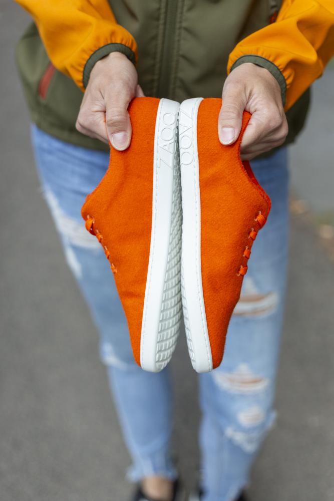 Eine Person hält ein Paar LIQE Orange-Sneaker von ZAQQ mit leuchtend orangefarbenem Obermaterial und weißer Sohle, die Sohle an Sohle vor einem verschwommenen Hintergrund präsentiert werden. Diese Sneaker versprechen ein außergewöhnliches Barfußgefühl. Die Person trägt eine grün-orangefarbene Jacke und blaue Distressed-Jeans.