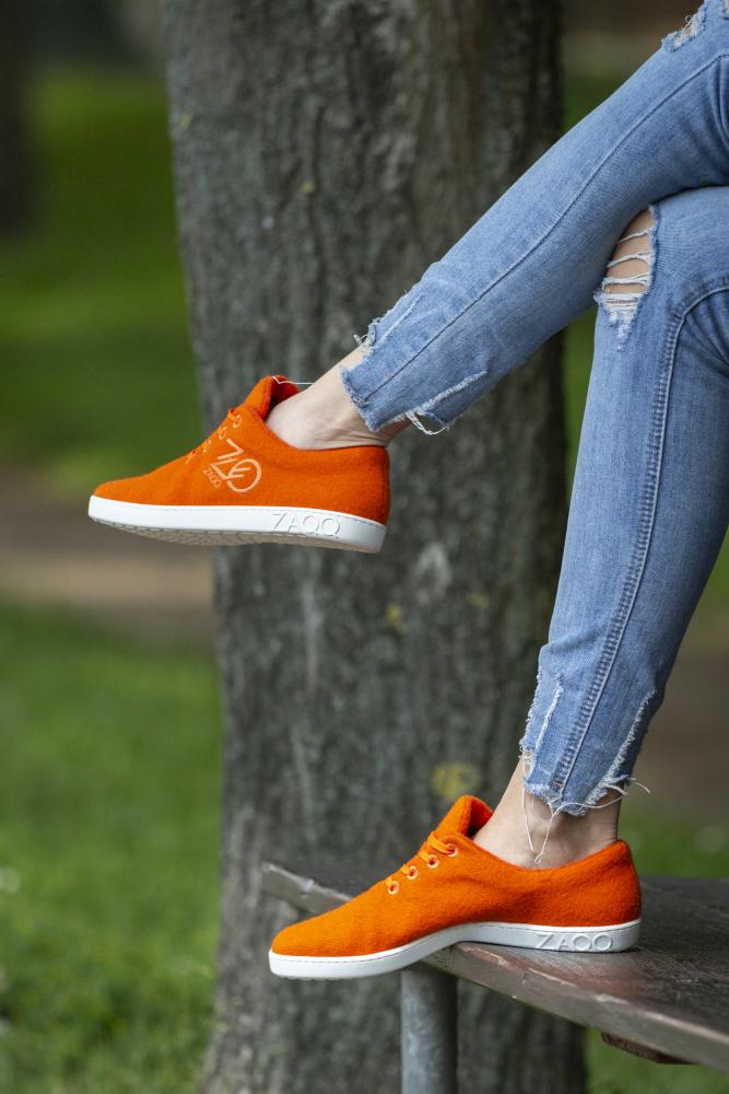 Eine Person, die die LIQE Orange Sneaker von ZAQQ mit ihrem auffälligen leuchtend orangefarbenen Design und den weißen Sohlen trägt, sitzt auf einer Bank und genießt den Barfußkomfort des Parks. Sie trägt zerrissene Bluejeans und sonnt sich unter Bäumen, die für eine natürliche Temperaturregulierung gegen die Mittagssonne sorgen.