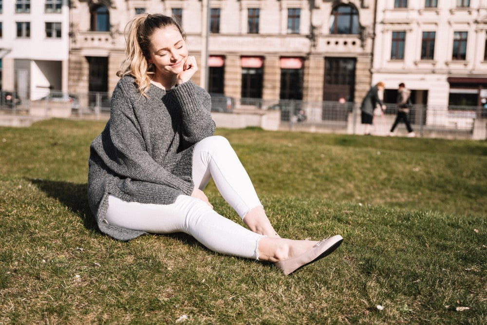 Eine Frau in einem LOOQ Nude-Pullover von ZAQQ und weißen Hosen sitzt anmutig im Gras, ihre Augen sind geschlossen und ihr Kopf ruht auf ihrer Hand. Sie strahlt Eleganz aus und wirkt entspannt mit einem historischen Gebäude im Hintergrund. Die Sonne scheint hell und lässt auf die Behaglichkeit eines warmen Tages schließen.