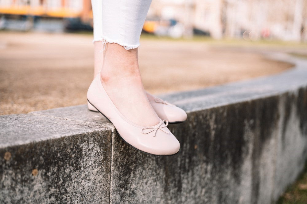 Nahaufnahme einer Person in weißen Jeans und ZAQQ’s LOOQ Nude Ballerinaschuhen mit eleganten Schleifen, die gemütlich auf einem Betonvorsprung vor einem unscharfen Parkhintergrund steht.