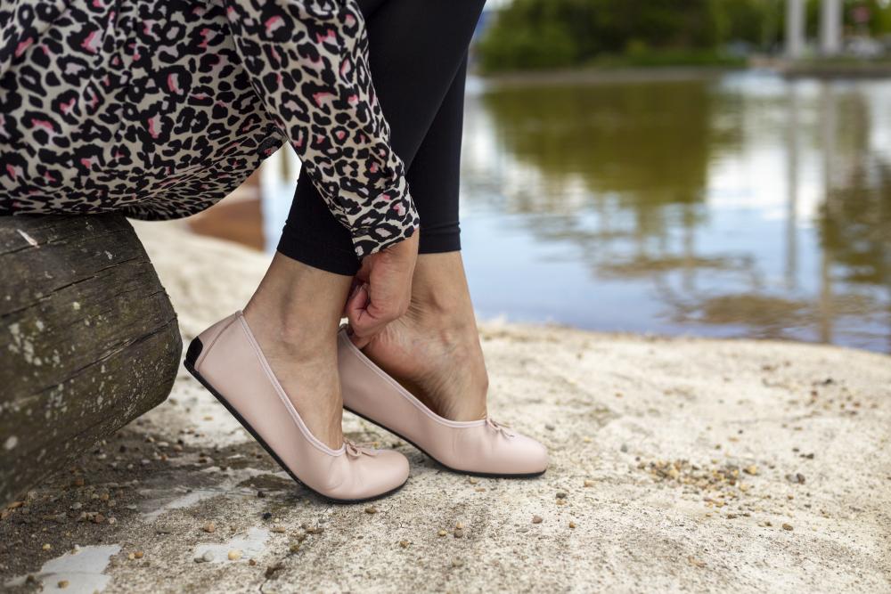 Eine Person sitzt auf einer Holzbank am Teich und rückt mit Eleganz ihre LOOQ Nude-Schuhe von ZAQQ zurecht. Sie trägt schwarze Leggings und ein Oberteil mit Leopardenmuster.