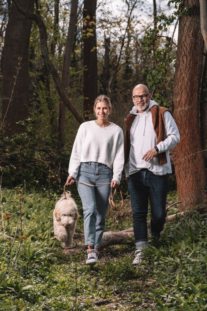 Ein Mann und eine Frau schlendern mit einem flauschigen Hund durch ein Waldstück. Die Frau, in einem weißen Pullover und Jeans, hält die Leine. Der Mann, der aus Bequemlichkeit die blau-weißen wasserdichten Schuhe ZAQQ MONTBLANQ trägt, lächelt, während sie gemeinsam ihren sonnigen Spaziergang genießen.