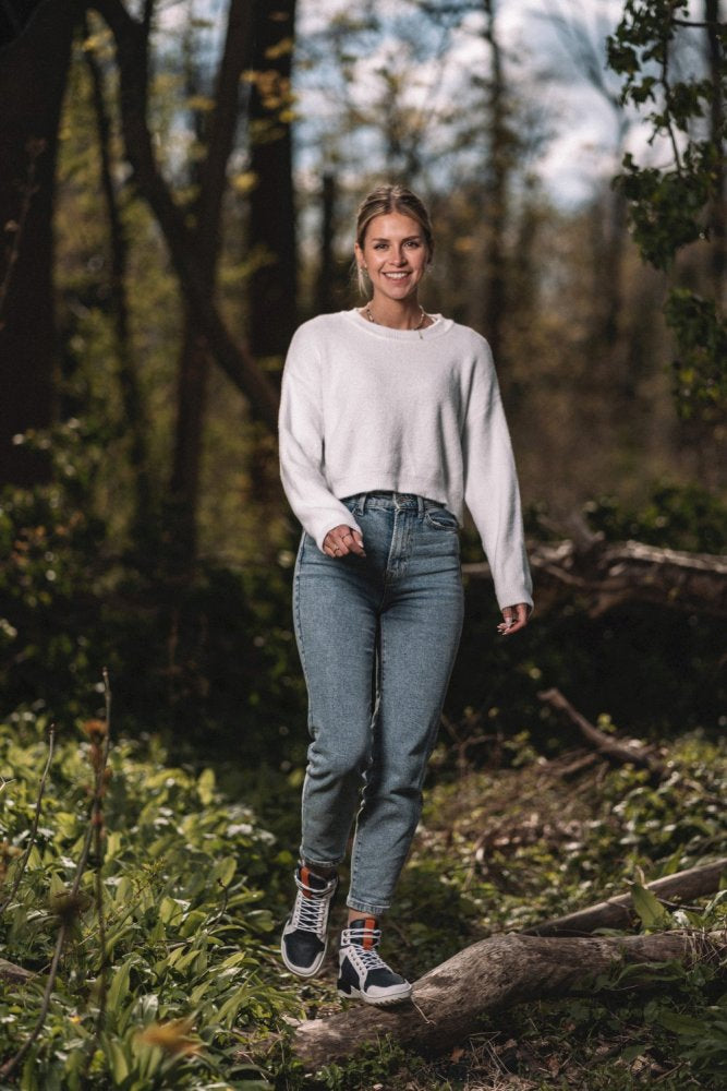 Eine Person in einem weißen Pullover und Jeans, die die stylischen blau-weißen wasserdichten Schuhe ZAQQ MONTBLANQ trägt, lächelt, während sie durch ein Waldstück geht. Sie steigt über einen umgestürzten Baumstamm inmitten von Bäumen und Grün und genießt den hellen und sonnigen Tag.