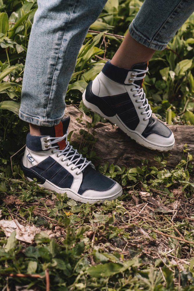 Eine Person, die die ZAQQ MONTBLANQ Blue White Waterproof High-Top-Sneaker trägt, steht mit einem Fuß auf einem Baumstamm, umgeben von Grün. Die hochgekrempelten hellblauen Jeans werten das lässige Outdoor-Ambiente auf und bringen diese vielseitigen Schuhe perfekt zur Geltung.