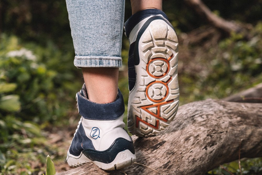 Nahaufnahme einer Person, die die blau-weißen wasserdichten Schuhe ZAQQ MONTBLANQ mit kräftigem orangefarbenem Schriftzug auf der TERRA-Sohle trägt und auf einem Baumstamm in einer Waldlandschaft steht. Die Schuhe sind teilweise sichtbar, die Jeans des Trägers ist hochgekrempelt.