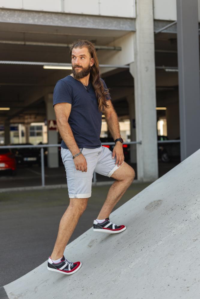 Ein Mann mit langen Haaren und Bart steht auf einer abschüssigen Betonfläche in einer städtischen Umgebung, gekleidet in ein marineblaues OAQLEY Red by ZAQQ T-Shirt, hellgraue Shorts und auffällige rot-schwarze Turnschuhe. Er blickt in die Ferne, einen Fuß selbstbewusst nach vorne gestellt.