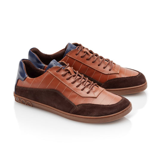 Wir präsentieren den QAMPION Brown von ZAQQ: Ein Paar stylische Sneaker aus elegantem braunem Leder und Wildleder, aufgewertet durch blaue Akzente an der Ferse. Diese Barfuß-Sneaker bestechen durch sorgfältige Nähte und sichere braune Schnürsenkel, alles auf einer flexiblen Kautschuksohle, die sich von einem strahlend weißen Hintergrund abhebt.