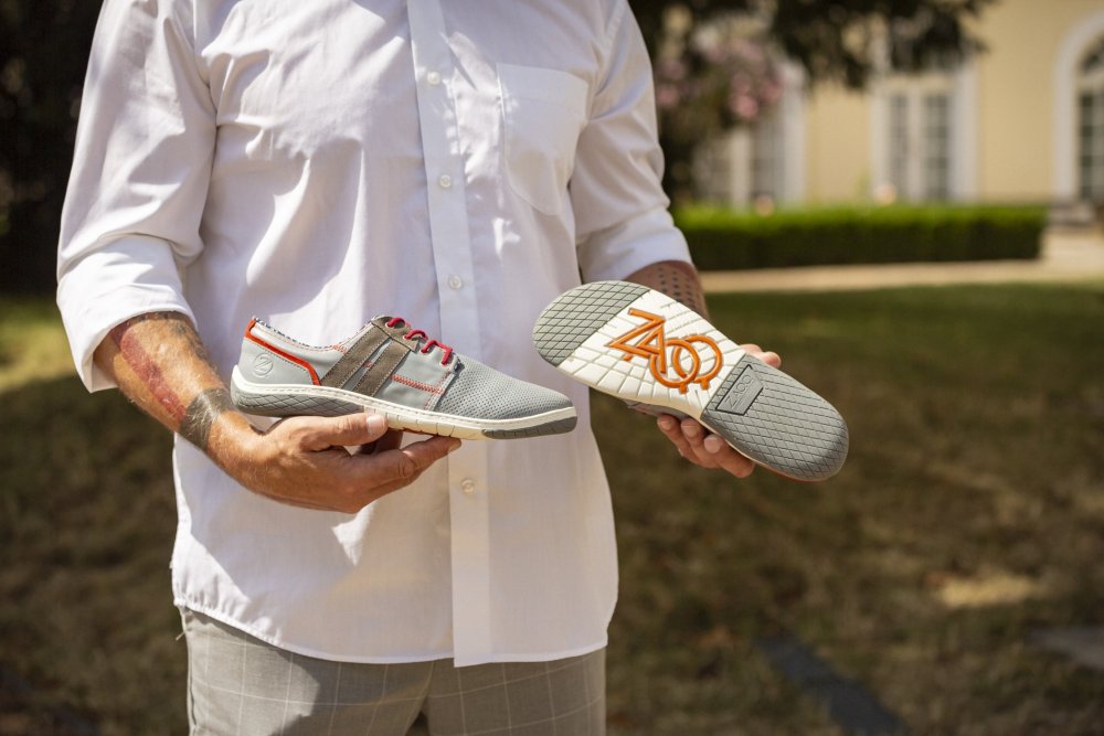 Eine Person in einem weißen Hemd hält ein Paar QANBERRA Grey-Schuhe von ZAQQ mit roten Akzenten. Ein Schuh hebt das Seitendesign hervor, während der andere die Sohle mit einem orangefarbenen Logo zeigt. Der Barfußschuh-Stil bildet einen schönen Kontrast zum Hintergrund aus Gras und einem Gebäude.