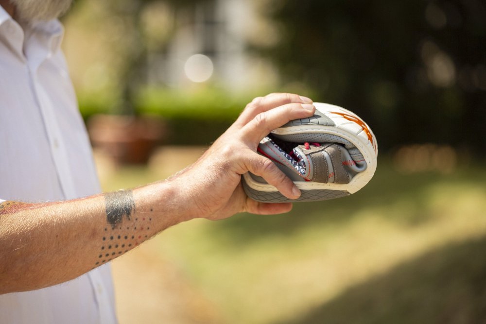 Eine Person mit tätowiertem Arm hält einen flexiblen ZAQQ QANBERRA Grey Barfußschuh, der mühelos in der Hand aufgerollt ist. Der Sneaker zeigt ein schlichtes Grau-Weiß-Farbschema, das durch auffällige rote Akzente ergänzt wird. Im unscharfen Hintergrund verstärken grüne Blätter und ein Gebäude die lebendige Szene.