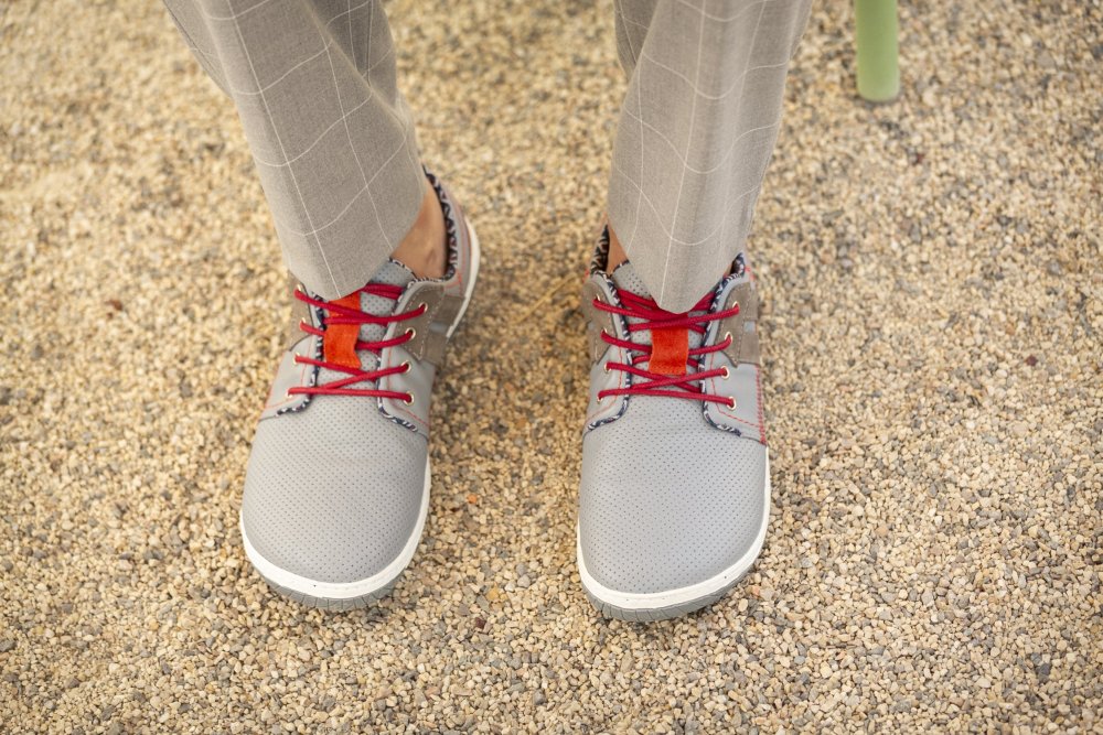 Ein Paar QANBERRA Grey-Schuhe von ZAQQ mit roten Schnürsenkeln und aus geschmeidigem Nappaleder gefertigt, wird von jemandem in hellgrauen karierten Hosen getragen, der barfuß auf einem Kiesboden steht.