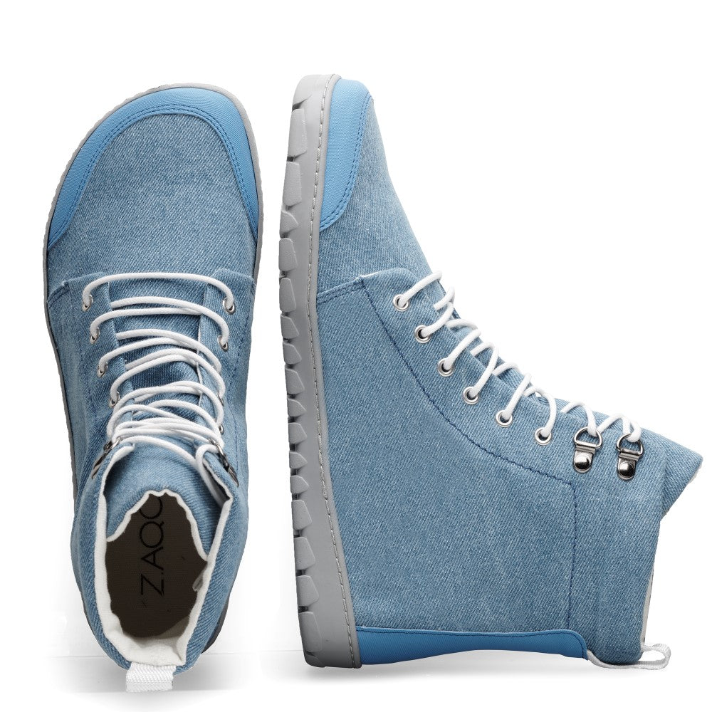 Ein Paar QICE Light Jeans-Sneaker von ZAQQ aus blauem Denim-Material mit weißen Schnürsenkeln und grauen Sohlen. Ein Schuh wird aufrecht präsentiert und zeigt die Seitenansicht und Sohle, während der andere flach liegt und die Oberseite und das Etikett auf der Innenseite zeigt, ideal für Liebhaber des Jeansstoff-Stils.