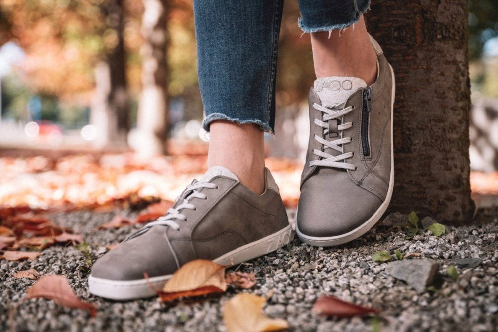 Eine Nahaufnahme einer Person, die ZAQQs QOMFY Wide Grey-Sneaker und Blue Jeans auf einem Kiesweg trägt. Überall verstreute Herbstblätter verstärken den Charme der Szene, während im Hintergrund ein Baum hervorlugt.