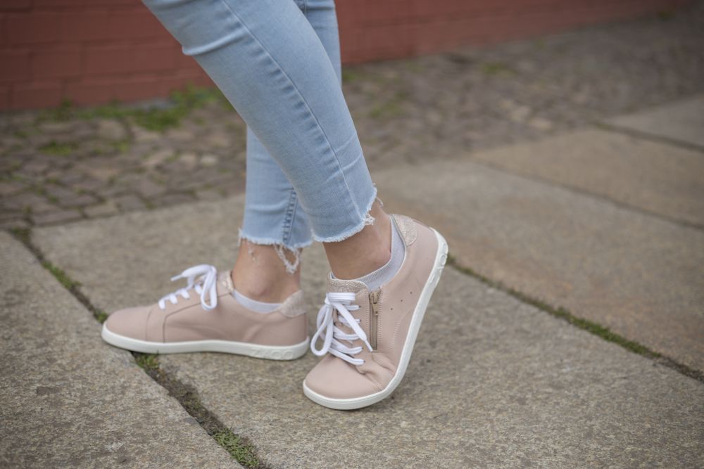 Eine Person in QOMFY Wide Rosa Jeans und beigefarbenen ZAQQ Sneakers aus hochwertigem Nappaleder mit weißer Sohle steht auf einem gepflasterten Weg. Die Jeans ist an den Knöcheln leicht ausgefranst, während eine rote Backsteinmauer einen lebendigen Hintergrund bildet.