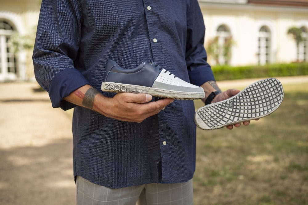 Eine Person in einem marineblauen Hemd steht im Freien und hält ein Paar QOOL Navy-Sneaker von ZAQQ. Ein Sneaker ist von der Seite zu sehen und zeigt sein Logo, während der andere mit der Sohle nach oben gehalten wird und ein speziell auf Komfort ausgelegtes Profil aufweist. Im Hintergrund sind Gebäude und Grünflächen zu sehen.