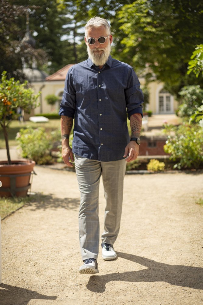 Ein bärtiger Mann mit Sonnenbrille, blauem Hemd und grauer Hose schreitet selbstbewusst einen sonnenbeschienenen Gartenweg entlang und genießt den Komfort seines ZAQQ QOOL Navy Barfuß-Sneakers. Im Hintergrund sind hohe Bäume und ein Gebäude zu sehen, Topfpflanzen säumen den Weg.