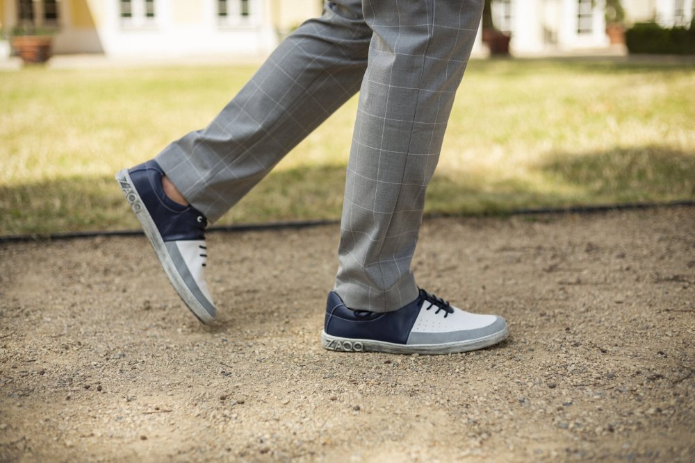 Eine Person in grau karierten Hosen und weißen ZAQQ QOOL Navy Sneakers mit blauen Akzenten schlendert einen Kiesweg entlang. Im Hintergrund sind Gras und unscharfe Gebäude zu sehen, was den außergewöhnlichen Tragekomfort des Schuhwerks unterstreicht.