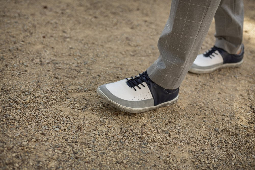 Eine Person in grau karierten Hosen und stylischen weißen ZAQQ QOOL Navy Sneakers schlendert auf einem Kiesweg und genießt den ultimativen Tragekomfort.