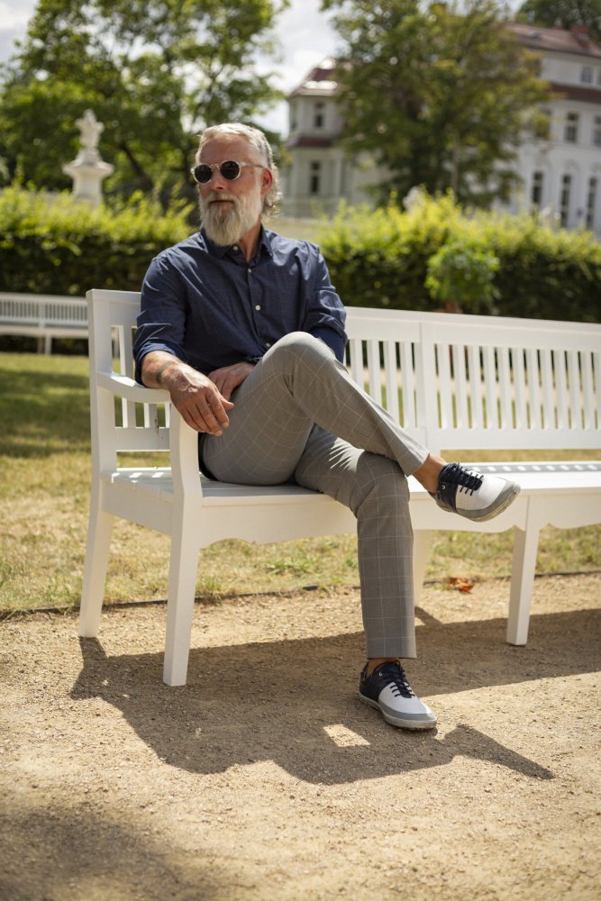 Ein Mann mit grauem Haar und Bart, einer Sonnenbrille, einem dunklen Hemd mit Knopfleiste, hellen karierten Hosen und den QOOL Navy-Sneakern von ZAQQ für zusätzlichen Komfort, sitzt auf einer weißen Bank in einem Garten. Das Sonnenlicht wirft weiche Schatten, im Hintergrund sind Bäume und ein Gebäude zu sehen.