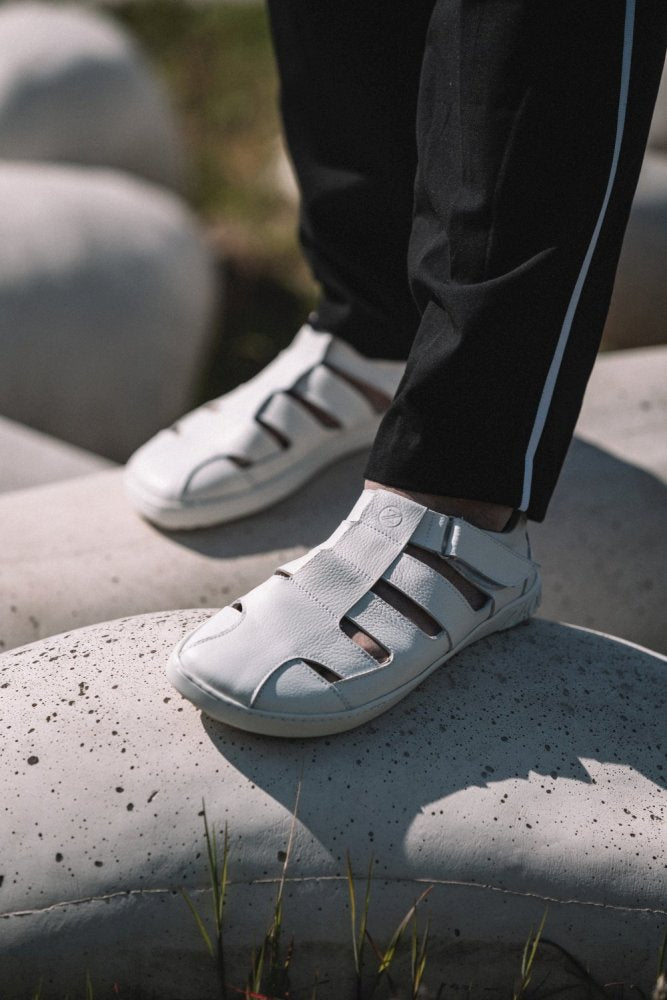 Eine Person in einer schwarzen Hose mit weißem Streifen und den QRUISE White Sandalen von ZAQQ steht im Freien auf großen runden Steinen und betont damit die Atmungsaktivität des Schuhwerks. Das Bild zeigt eine Nahaufnahme der Füße und Beine.